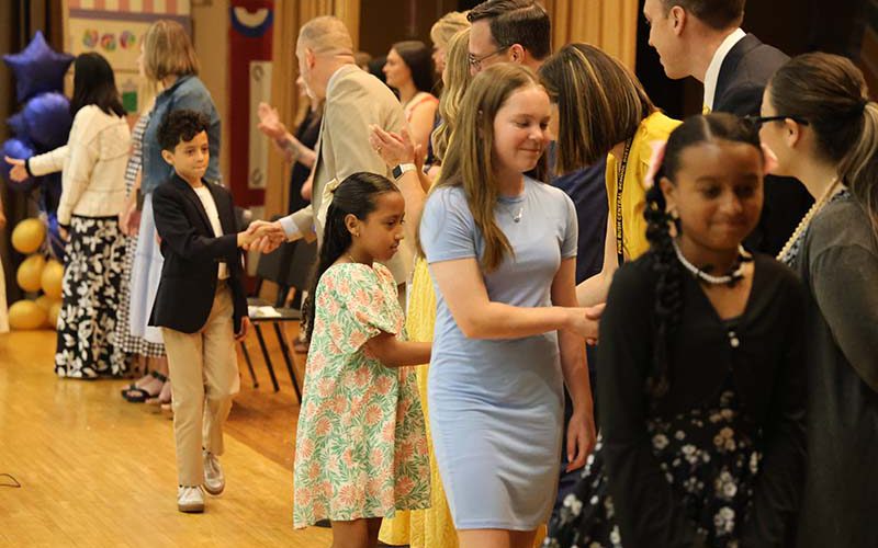 A line of fifth-grade students shake hands with adults as they walk across a stage.