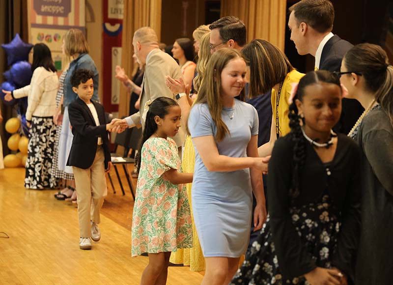 A line of fifth-grade students shake hands with adults as they walk across a stage.