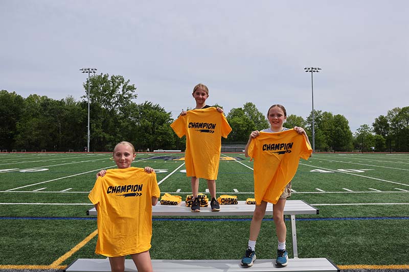 Three fourth-grade girls stand on a podium, each holdin gup a yellow tshirt that says champion.