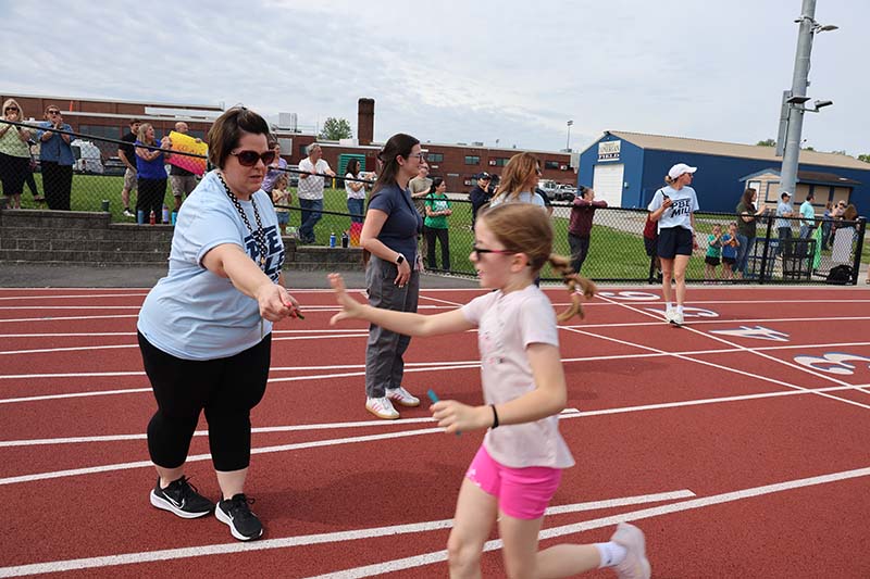 An elementary age girl takes a popsicle stick from an adult as she runs on a track.