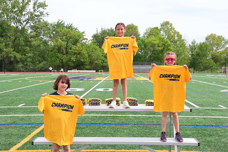 Three third-grade girls stand on a podium, each holdin gup a yellow tshirt that says champion.