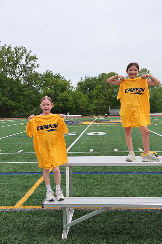 Two fifth-grade girls hold up yellow tshirts that say champion.