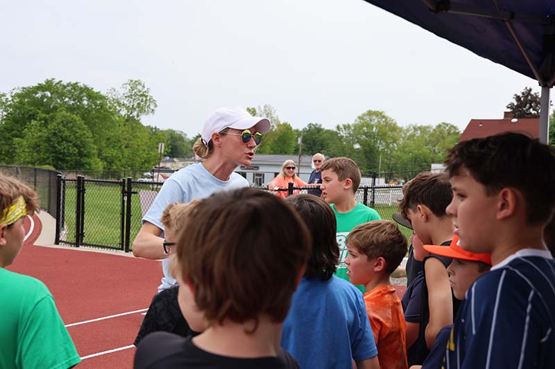 A woman wearing a white cap stands in the midst of elementary boys talking to them on a track.