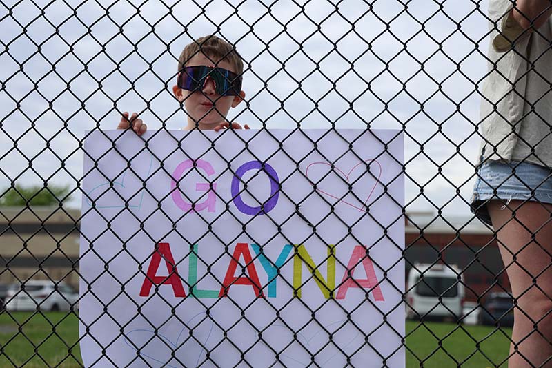 A kid wearing sunglasses stands behind a fence holding a sign that says Go Alayna.