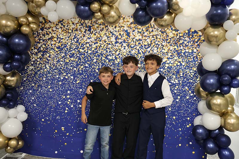 Three fifth-grade boys stand in front of a blue and gold sparkly backdrop with blue white and gold balloons around it.