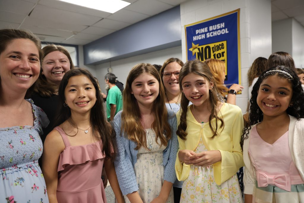 A woman on the left stands with a group of fifth-grade girls, all are smiling.