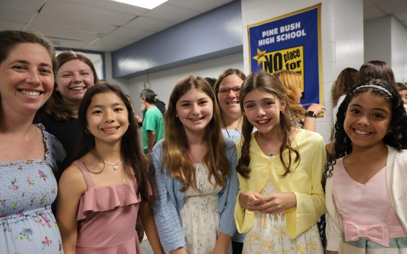 A woman on the left stands with a group of fifth-grade girls, all are smiling.