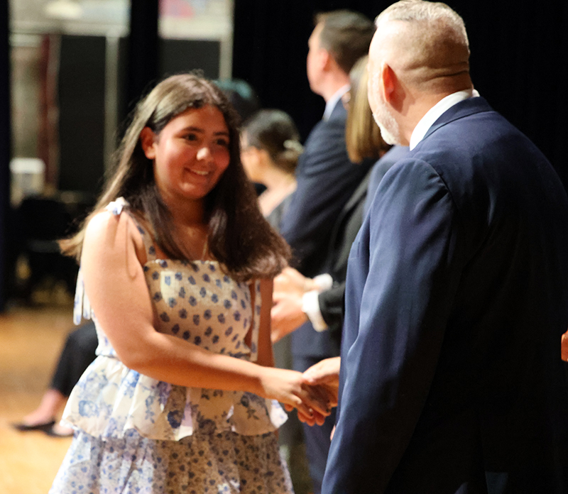 A fifth-grade girl in a polkadot dress shakes hands with a man in a suit.