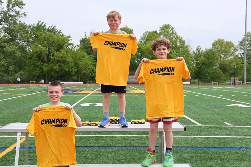 Three fourth-grade boys stand on a podium, each holdin gup a yellow tshirt that says champion.