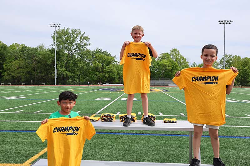 Three third-grade boys stand on a podium, each holdin gup a yellow tshirt that says champion.