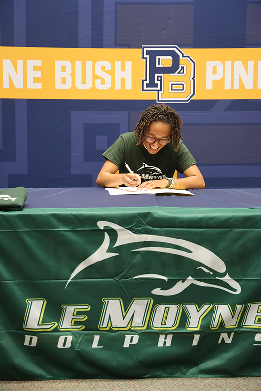 A high school senior smiles as she writes on a piece of paper. Behind her is a Pine Bush banner and the table cover says LeMoyne Dolphins.