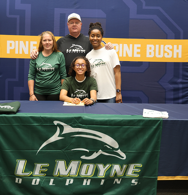 Three adults stand behind a young woman who is sitting after signing her national letter of intent to run track in college. The table cover says LeMoyne Dolphins.
