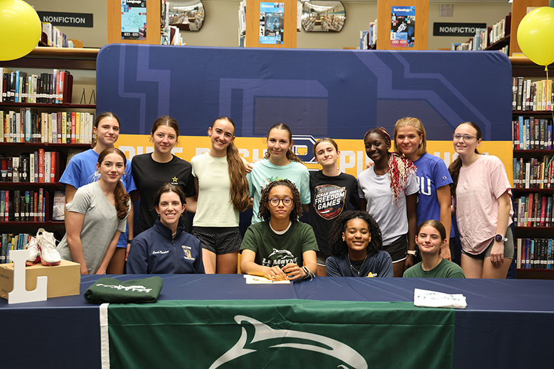 A group of 12 high school girls stand around a high school senior who has just signed her letter of intent to run track at LeMoyne College.