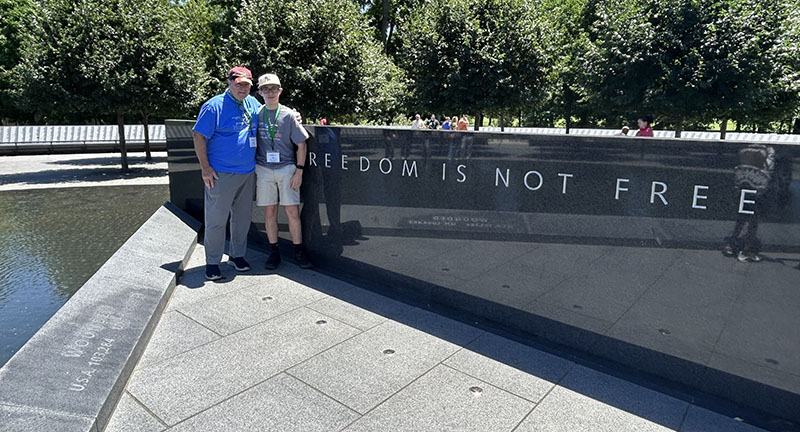 A man in a blue shirt and baseball cap stands with a young man with glasses and a baseball cap in front of a wall that says freedom is not free.