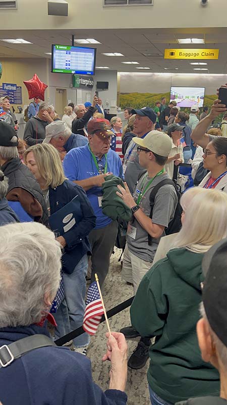 A man in a blue shirt and baseball cap stands with a young man with glasses and a baseball cap stand among a crowd of people.