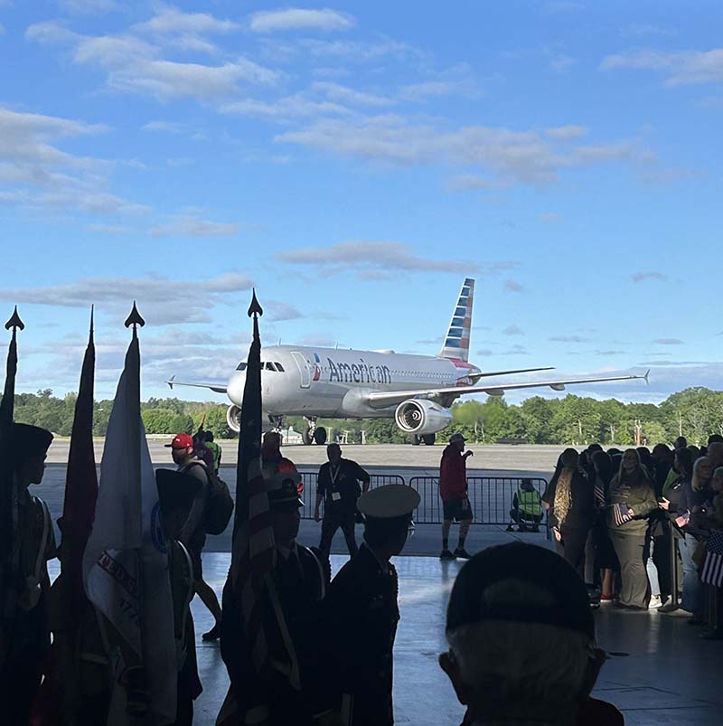An American Airlines airplane is on the tarmac. Sky is blue. There are people holding flags in front.