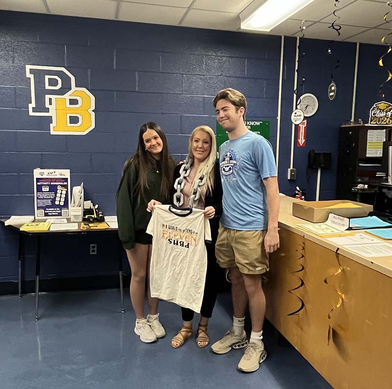 A woman stands between two high school students. She is holding a white tshirt. All are smiling.