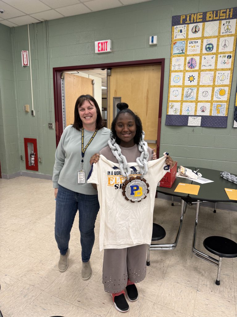 A high school student smiles as she holds up a white tshirt and is wearing a huge chain with a huge medallion that says PB. She is standing with a woman who is smiling.
