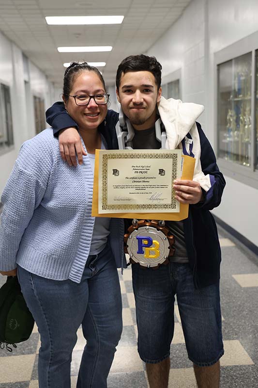 A woman in a blue sweater smiles as she stands with a young man holding a certificate.