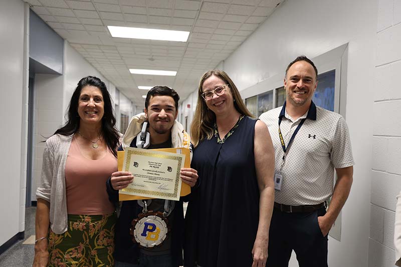 Three adults stand with a high school senior. All are smiling. The student holds up a certificate and t shirt and is wearing a huge chunky chain with a big PB medallion.
