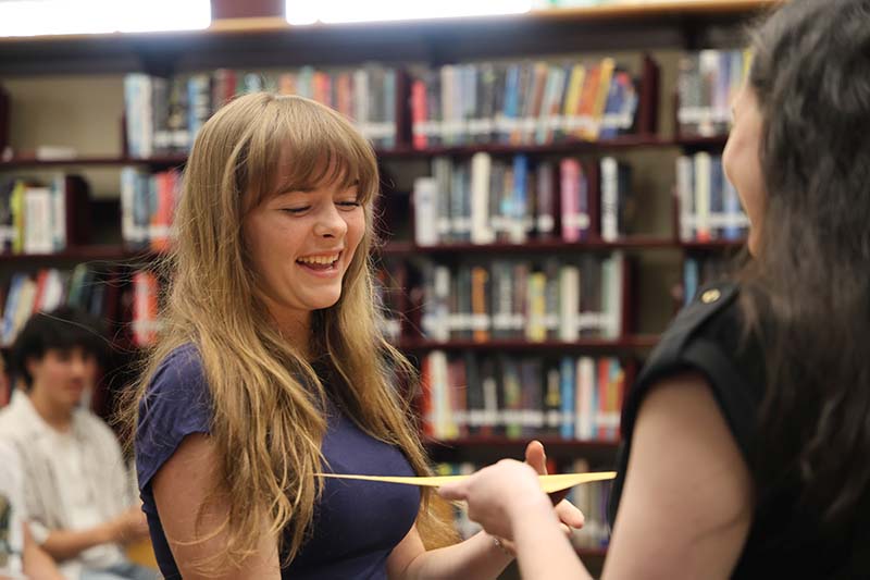 A high school girl with long blonde hair shakes hands with a woman and accepts a large envelope from her.