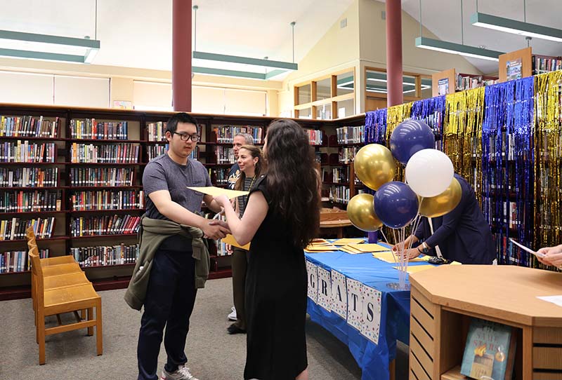 A high school boy shakes hands with a woman and accepts a large envelope from her.