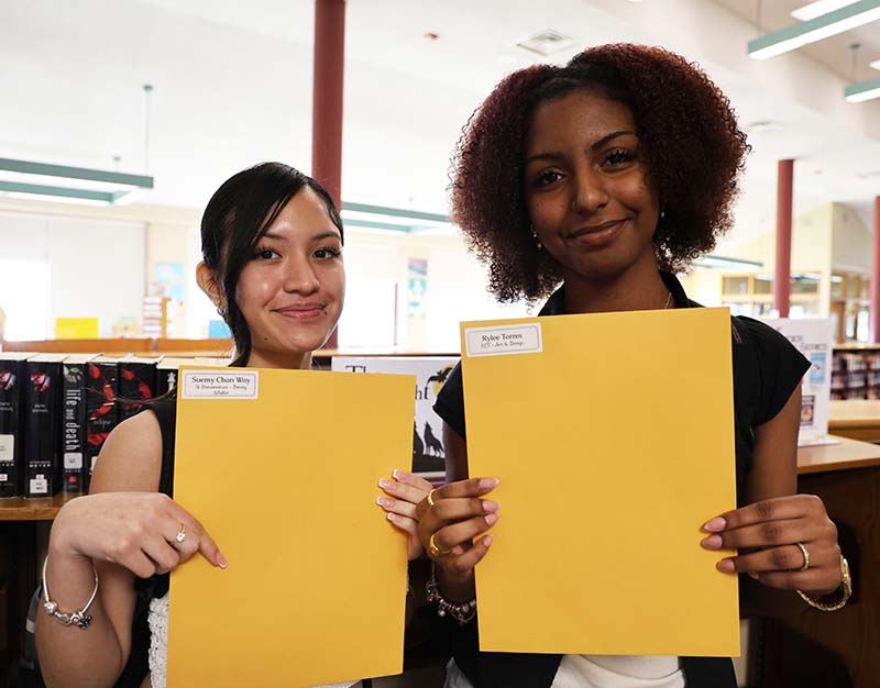 Two high school girls smile and hold up large envelopes they received.