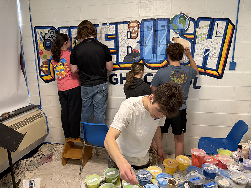 Several artists paint parts of a large mural on a wall that says Pine Bush High School.