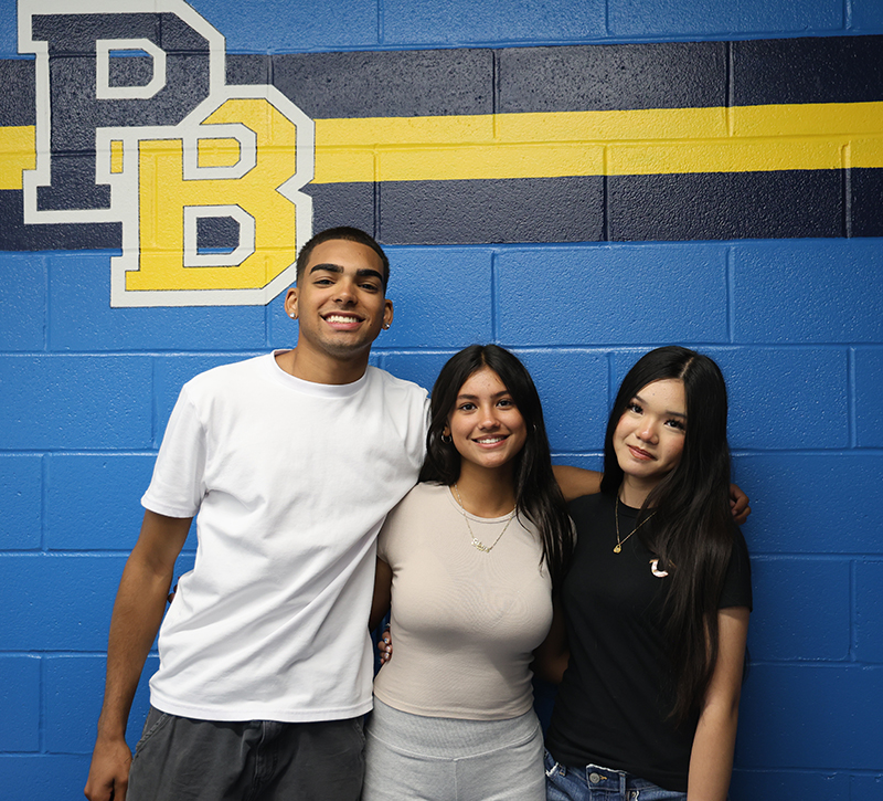 A blue wall background with a large PB painted near the top with blue and gold stripes. Three high school students stand together, a young man and two young women.