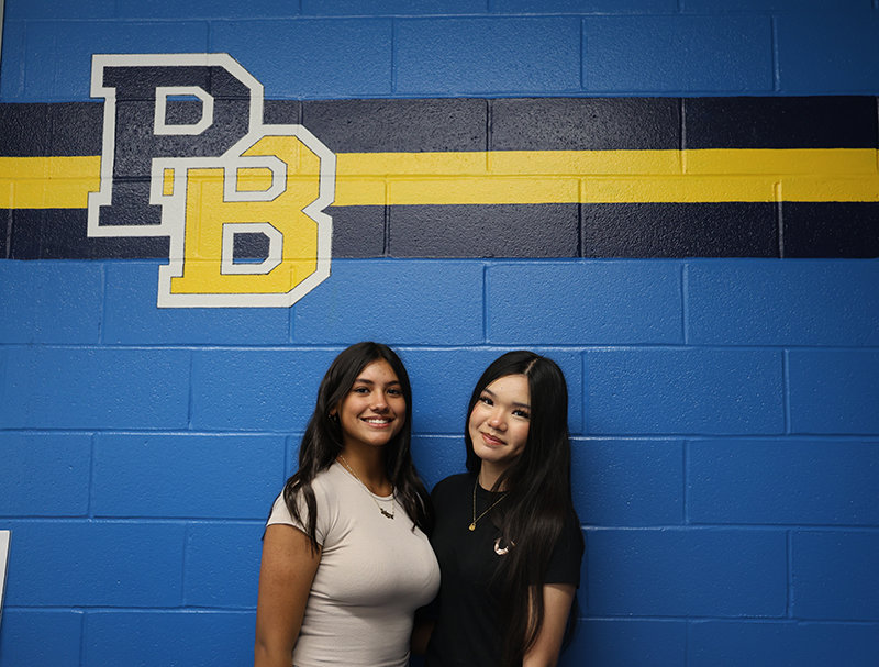 A blue wall background with a large PB painted near the top with blue and gold stripes. Two young women, high school students, stand together and smile.