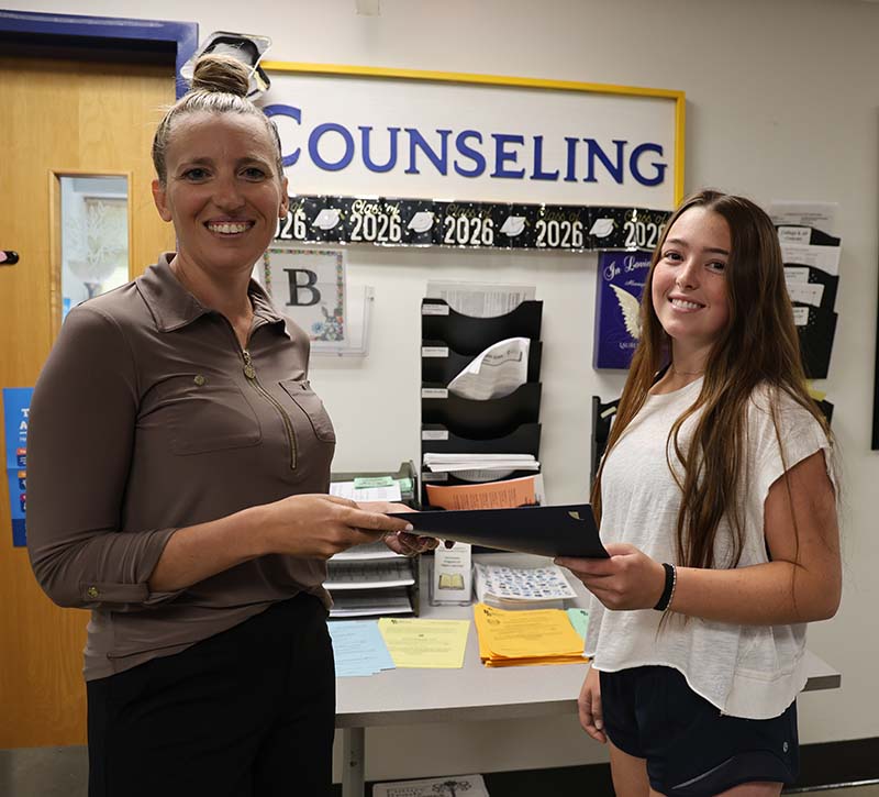 A woman on the left smiles as she hands an award to a high school junior girl who is also smiling.
