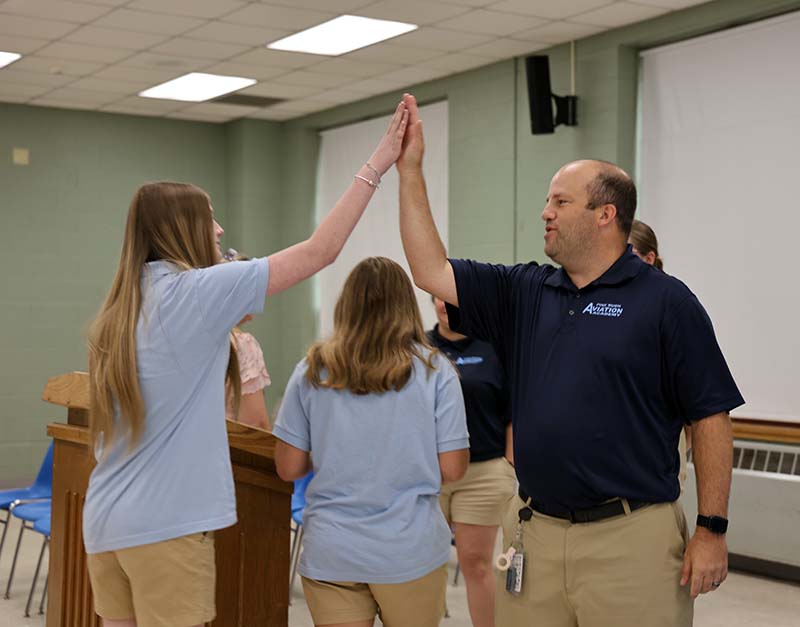 A man in a dark blue polo shirt high fives a high school student with long blonde hair.