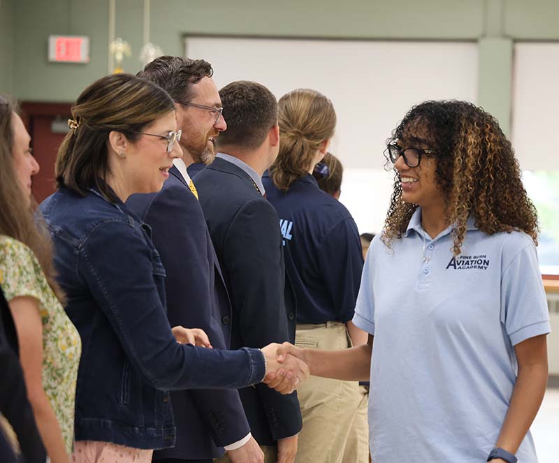 A high school girl with long darker hair smiles and shakes hands with a woman.