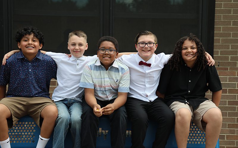 Five fifth-grade boys sit on a bench, all dressed up, with their arms around each other.
