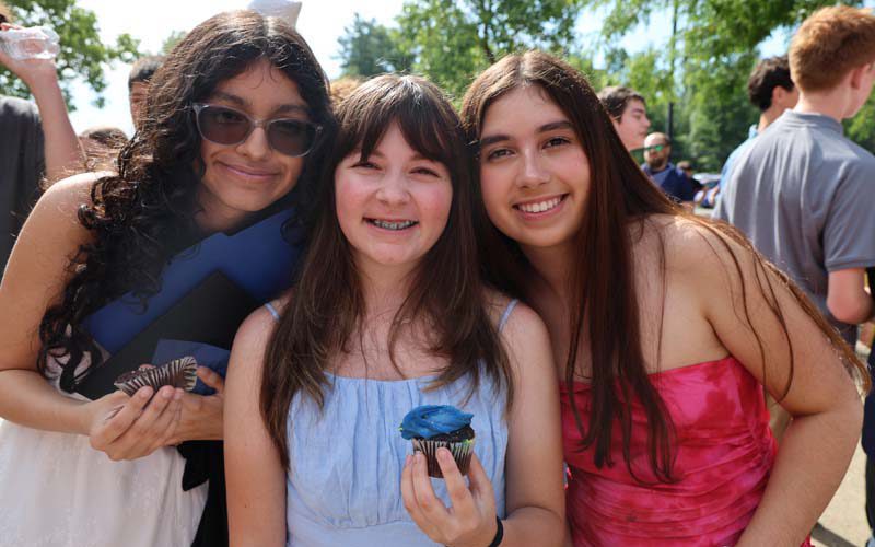 Three eighth-grade girls smile. One is holding a blue cupcake.