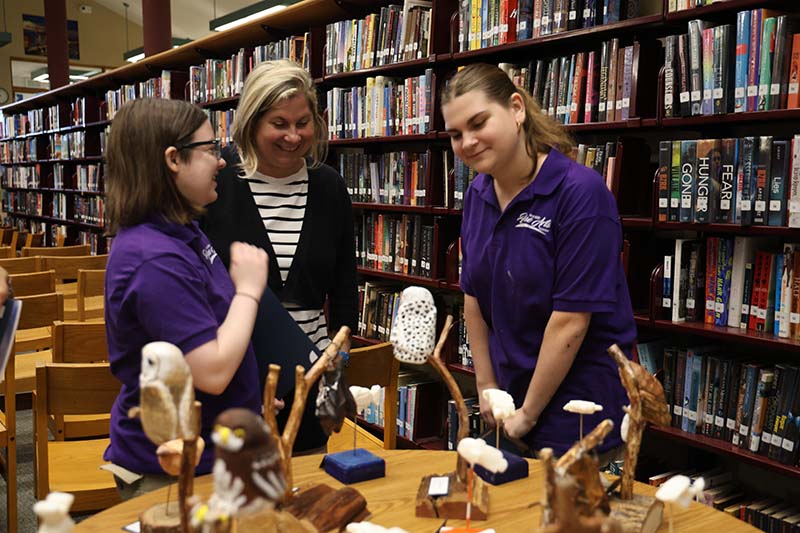 A woman and two high school students stand around a table looking at artwork.