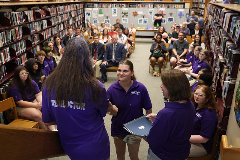 A young woman shakes hands with a woman. Behind the young woman are people sitting on chairs.