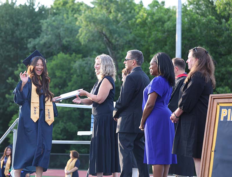 A high school senior dressed in a blue cap and gown with a gold sash around her shoulders smiles as she accepts her diploma from a woman, who is surrounded by other adults on a stage outdoors.