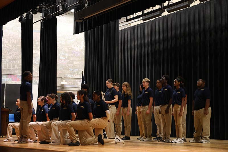 A group of high school students all wearing blue shirts and khaki pants stand on a stage in a formation. One has a drum.