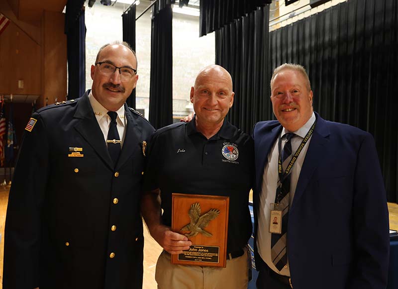 Three men stand on a stage. The one in the middle holds a plaque.