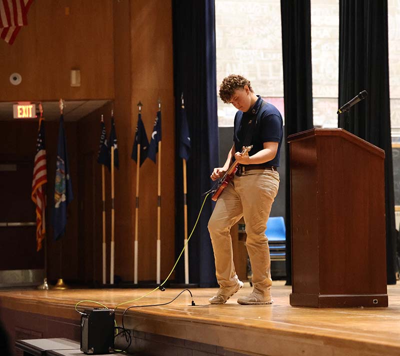 A high school boy, wearing a blue polo shirt and khaki pants plays the electric guitar on a stage.