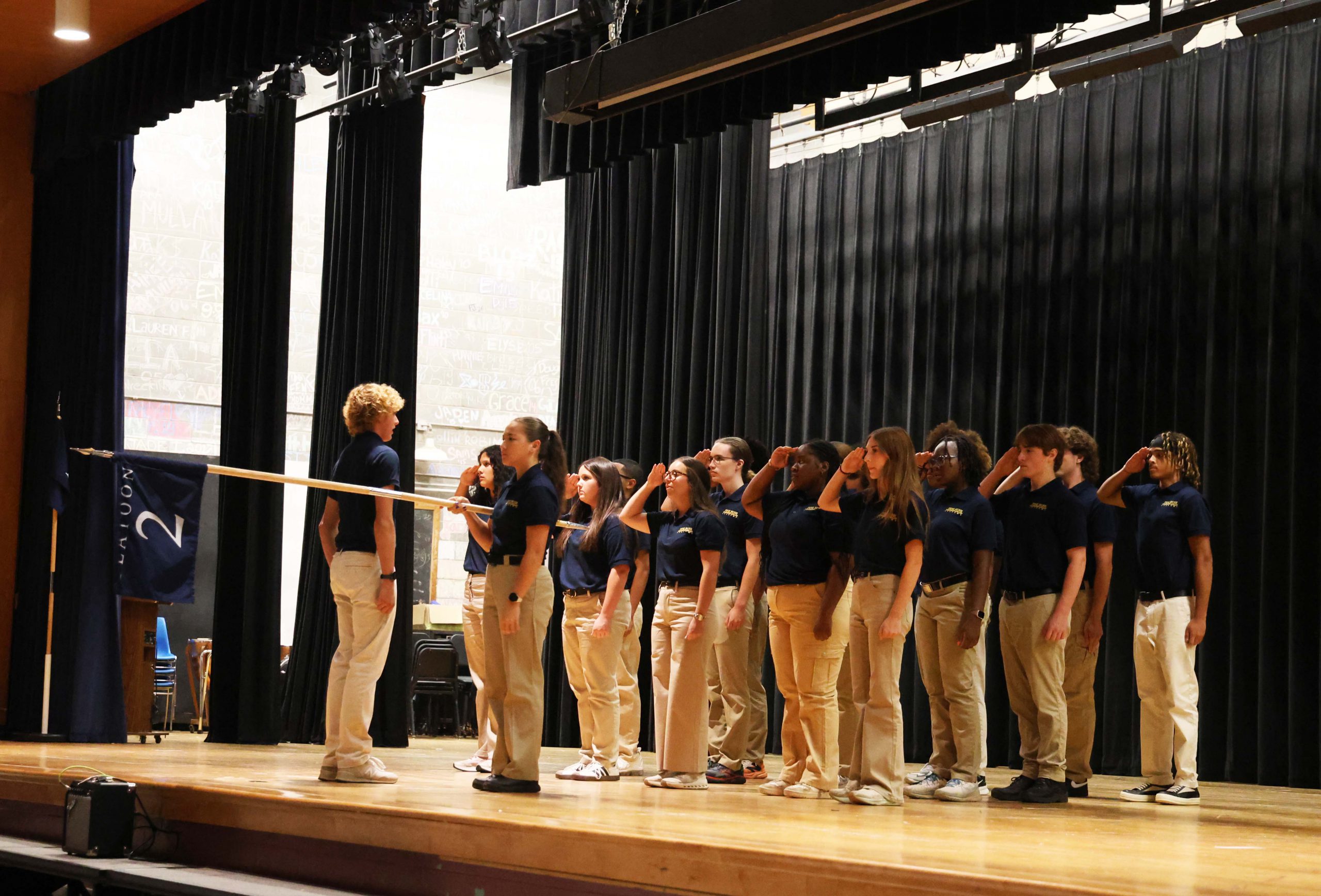 A group of high school kids all wearing blue polo shirts and khaki pants stand on a stage in a formation saluting. One is holding a flag straight out.