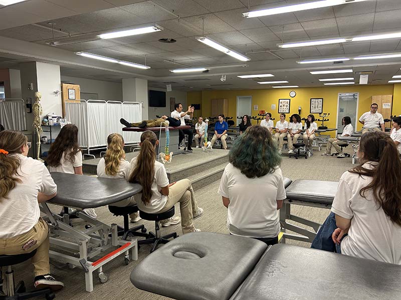 A group of high school students sit around a large room listening to an instructor.