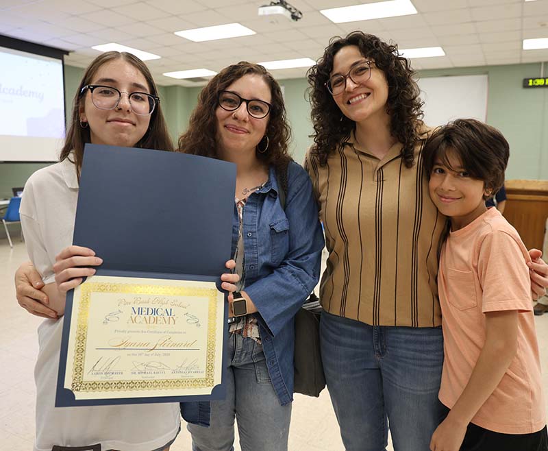 Two adults stand with a young woman who is holding a certificate. There is a younger child with them. all are smiling.