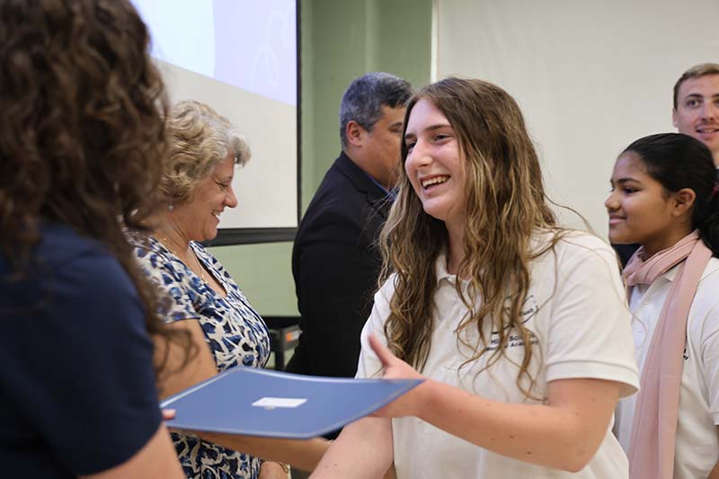 A young woman with long dark hair smiles and shakes hands with someone who hands her a certificate.