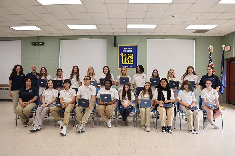 Twenty two high school students, all wearing khaki pants and white polo shirts sit and stand holding certificates. There are three adults standing on the sides.