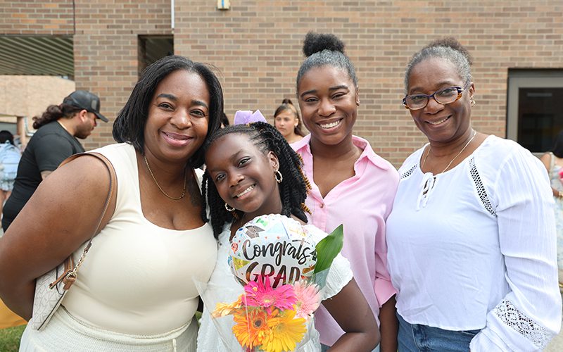 A fifth-grade girl holding flowers smiles as she stands with three women.