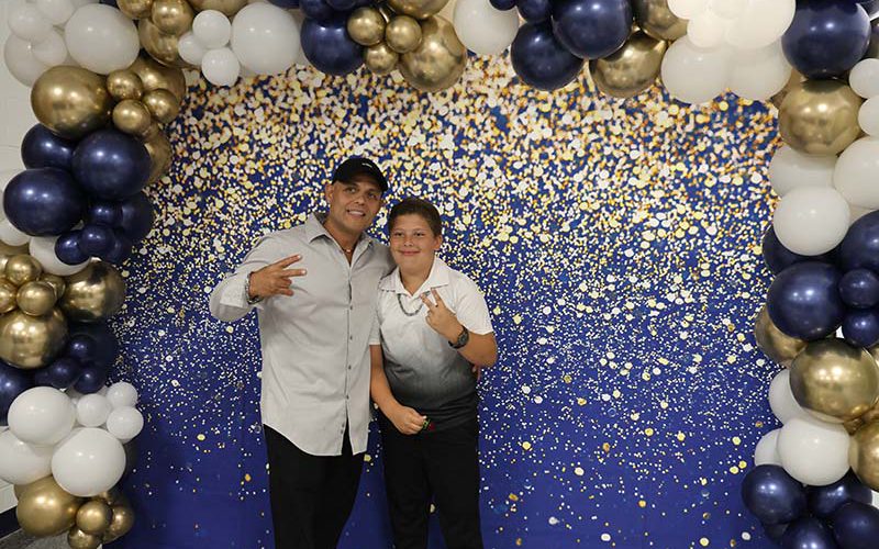 Two fifth-grade boys give thumbs up under an arch of blue, gold and white balloons.