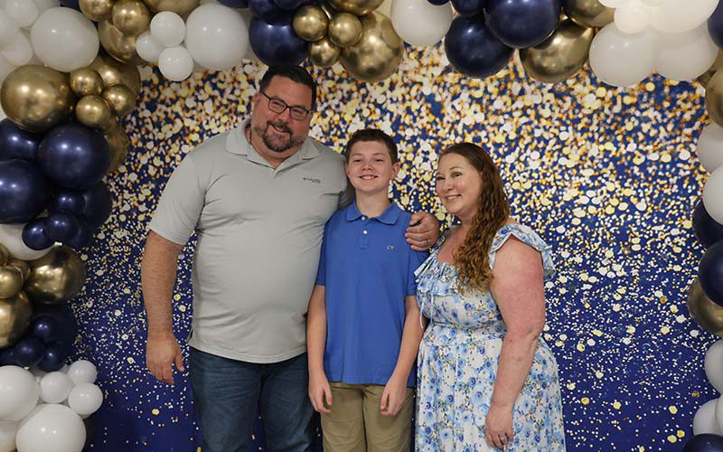 A fifth-grade boy stands in between a man and a woman, smiling, under a blue, white and gold balloon arch.