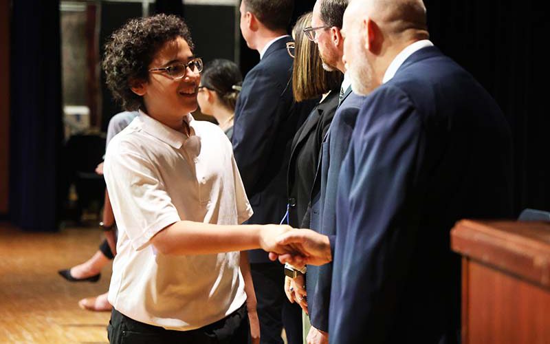 A fifth-grade boy shakes hands with a man on a stage.